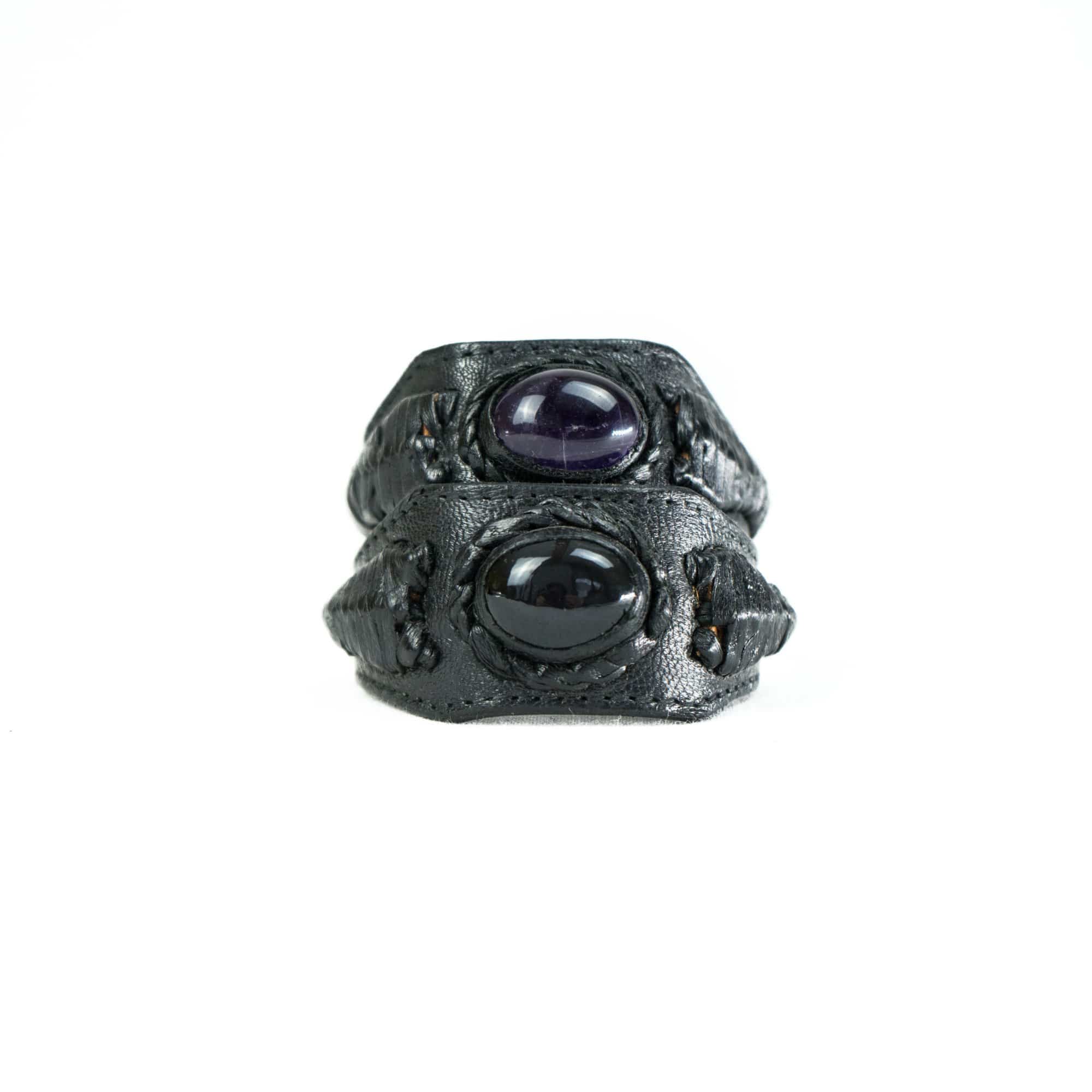 A close-up of the ANOM Men's Black Cuff Leather Bracelet by Kaya Movement, featuring an oval-shaped, dark purple genuine Amethyst gemstone and a round, black stone, with intricate textured detailing along the band, set against a plain white background.